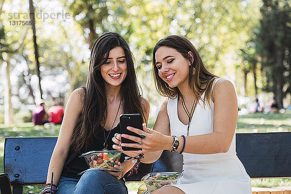 Freundinnen sitzen auf einer Parkbank und schauen auf ein Smartphone