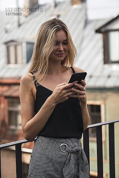 Porträt einer lächelnden blonden Frau  die auf dem Balkon steht und auf ihr Handy schaut