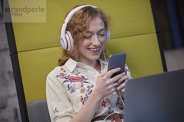 Junge Frau  die in einem modernen Kreativbüro arbeitet  Kopfhörer trägt und ein Smartphone benutzt
