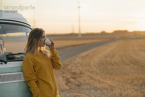 Junge Frau trinkt bei Sonnenuntergang aus Becher am Wohnmobil in ländlicher Landschaft