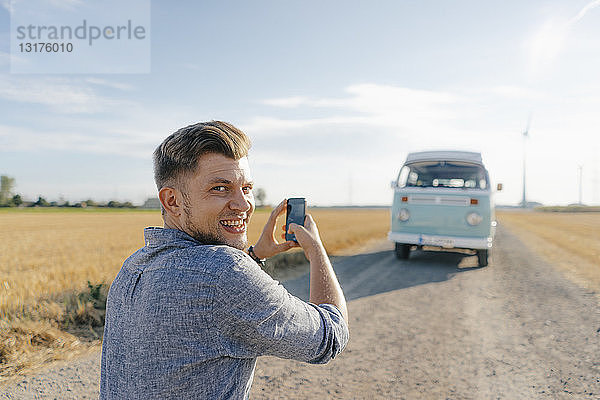 Lächelnder junger Mann beim Fotografieren eines Wohnmobils in ländlicher Umgebung mit dem Handy