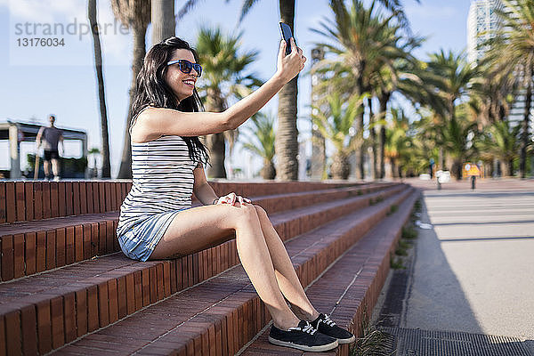 Spanien  Barcelona  junge Frau sitzt auf einer Treppe im Sonnenlicht und macht Selbsthilfe mit einem Smartphone