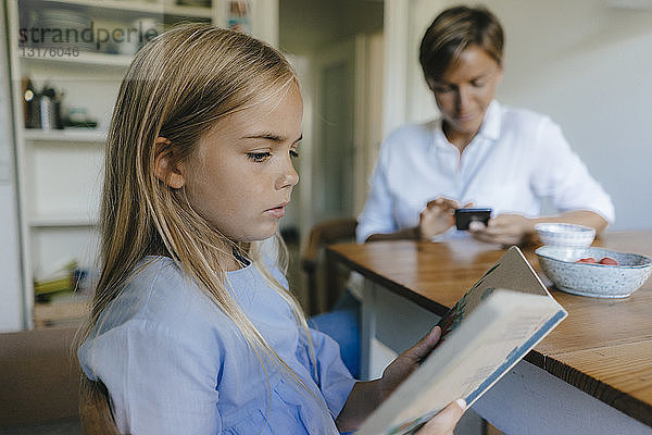 Mädchen mit Buch sitzt zu Hause mit der Mutter am Tisch und benutzt im Hintergrund ein Handy