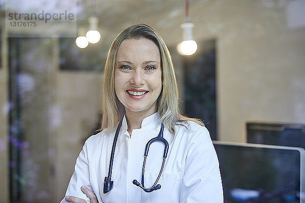 Porträt einer lächelnden Ärztin mit Stethoskop hinter Fensterscheibe