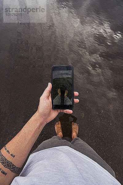 Mann macht ein Handyfoto mit Füßen im Wasser eines Sees
