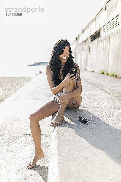 Lächelnde junge Frau mit Nasenpiercing und Tätowierung mit Smartphone in Strandnähe