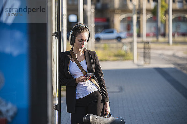 Junge Frau mit Kopfhörern und Handy an der Straßenbahnhaltestelle