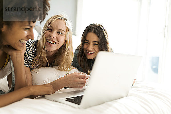 Drei glückliche Freundinnen liegen im Bett und teilen sich einen Laptop