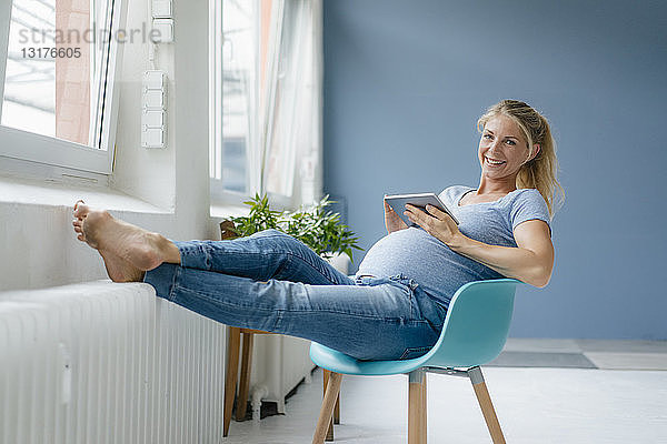 Porträt einer lächelnden schwangeren Frau  die auf einem Stuhl am Fenster sitzt und ein Tablett hält