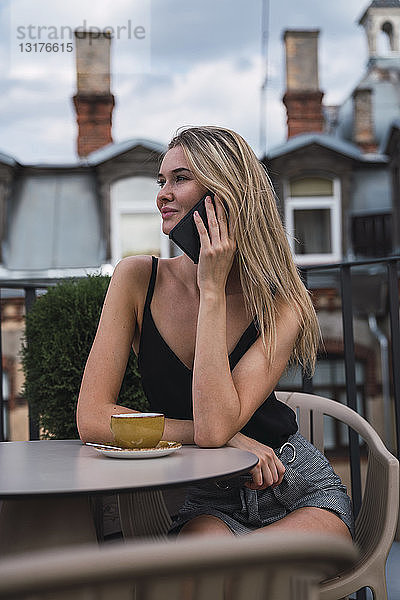 Lächelnde blonde Frau am Telefon  die auf der Dachterrasse bei einer Tasse Kaffee sitzt