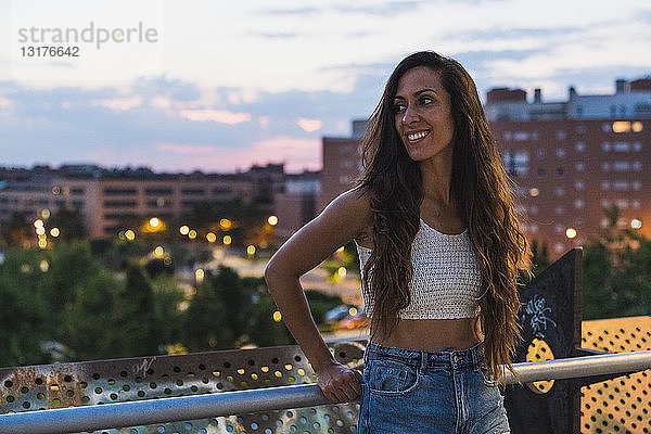 Schöne lächelnde junge Frau mit langen braunen Haaren in der Stadt in der Abenddämmerung