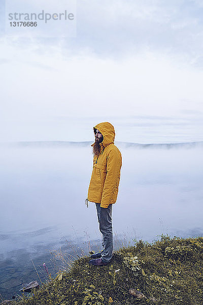 Schweden  Lappland  Mann mit Vollbart und gelber Windjacke steht am Wasser und schaut in die Ferne