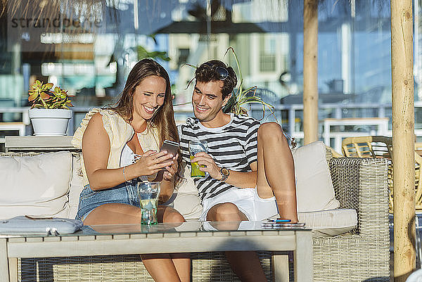 Glückliches junges Paar sitzt auf der Terrasse einer Bar und schaut auf sein Handy