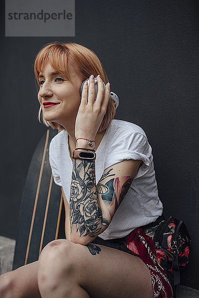 Lächelnde junge Frau mit Skateboard und Kopfhörern beim Musikhören