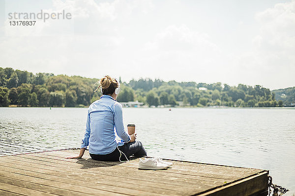 Frau sitzt mit Kopfhörern und Kaffee zum Mitnehmen auf einem Steg an einem See