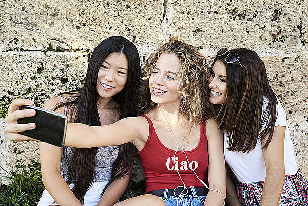 Drei lächelnde junge Frauen sitzen an einer Steinmauer und machen ein Selfie