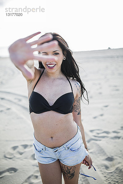 Porträt einer lächelnden jungen Frau mit Nasenpiercing und Tätowierung  die am Strand durch ihre Finger blickt