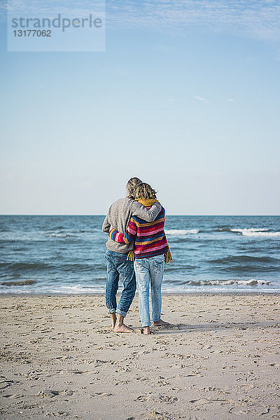 Ein Paar  das mit den Armen am Strand steht und auf das Meer schaut