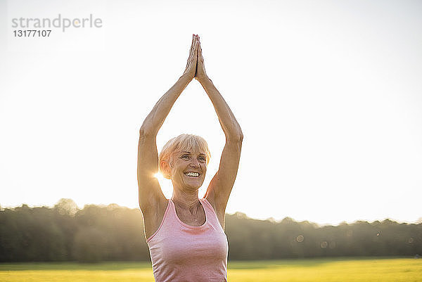 Lächelnde ältere Frau macht Yoga auf einer ländlichen Wiese bei Sonnenuntergang