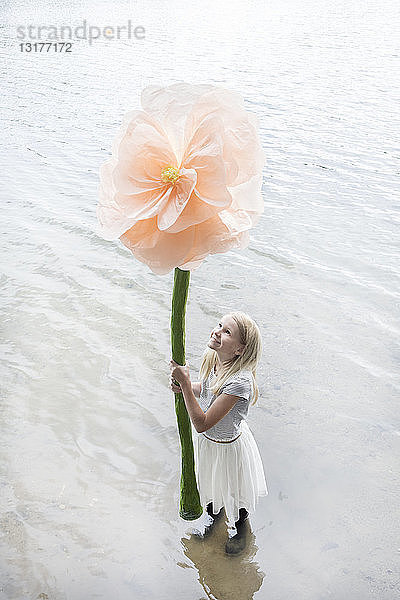 Lächelndes blondes Mädchen steht in einem See und hält eine überdimensionale Kunstblume