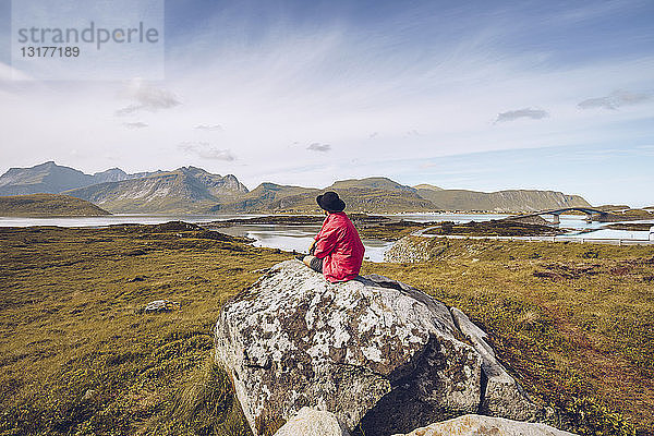 Norwegen  Lofoten  Mann in roter Regenjacke sitzt auf einem Felsen und schaut auf die Aussicht