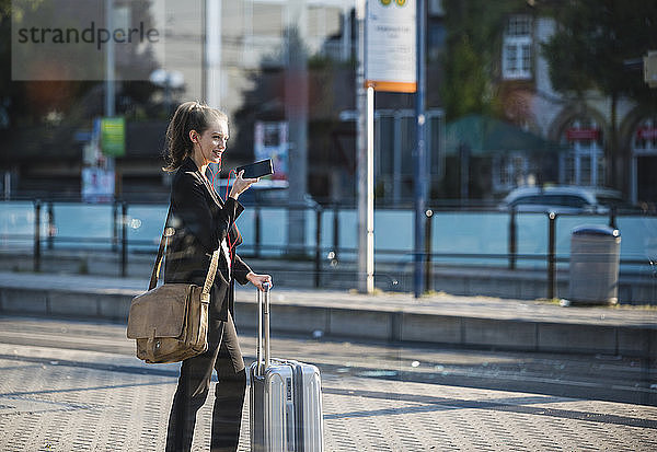 Lächelnde junge Frau mit Gepäck an der Straßenbahnhaltestelle in der Stadt per Handy