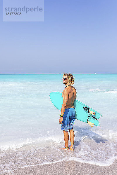 Junger Mann steht am Meer und hält sein Surfbrett
