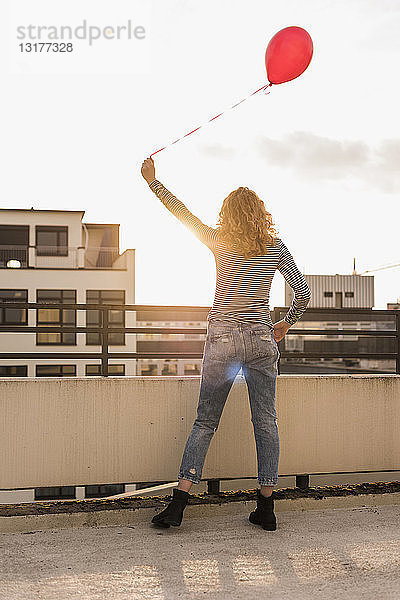 Rückenansicht eines jungen Mannes mit rotem Ballon  der bei Sonnenuntergang auf der Dachterrasse steht