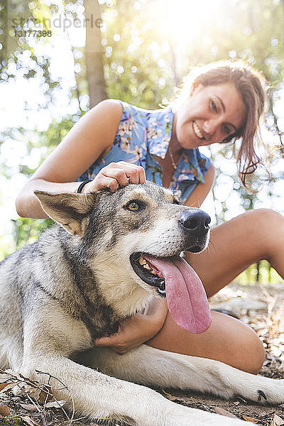 Lächelnde junge Frau sitzt auf dem Waldboden und streichelt ihren Hund