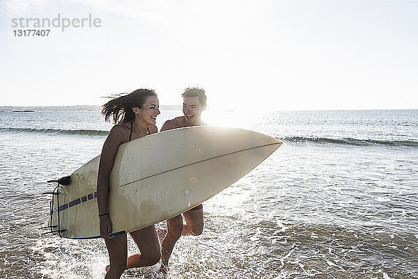 Frankreich  Bretagne  glückliches junges Paar mit im Meer laufendem Surfbrett