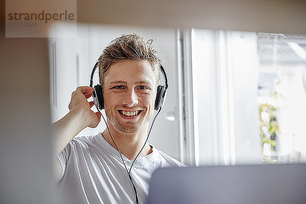 Porträt eines lächelnden jungen Mannes zu Hause  der mit Kopfhörern Musik hört