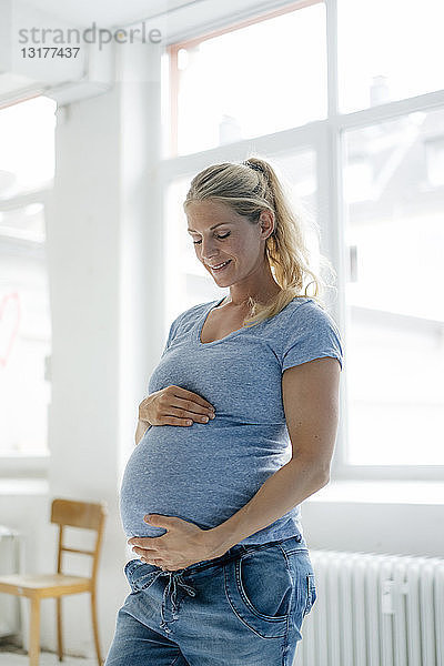 Lächelnde schwangere Frau steht am Fenster und schaut auf ihren Bauch