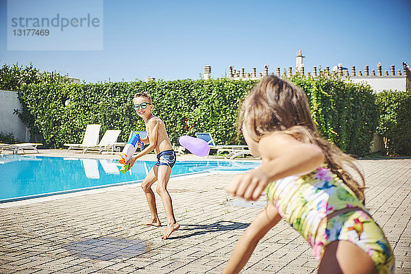 Mädchen und Junge haben eine Wasserschlacht mit Wasserpistole und Wasserbomben am Beckenrand