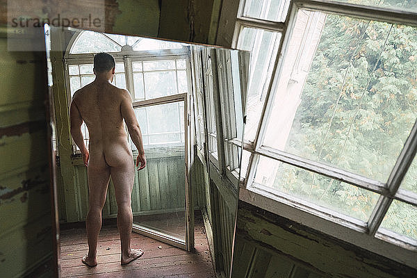 Spiegelbild eines nackten Mannes  der am Fenster eines verlassenen Hauses steht