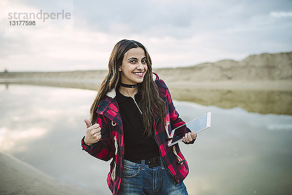 Porträt einer lächelnden jungen Frau  die sich am Strand mit einem Smartphone selbstständig macht