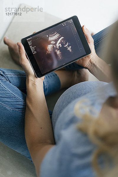Schwangere Frau sitzt auf dem Boden und betrachtet Ultraschallbild auf Tablette