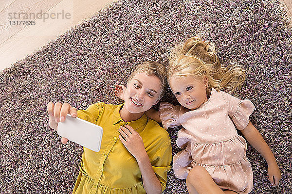 Lächelnde Mutter und Tochter auf dem Teppich liegend  die ein Selfie