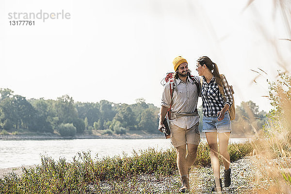 Glückliches junges Paar mit Rucksäcken und Gitarre beim Spaziergang am Flussufer