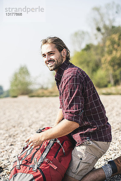 Lächelnder junger Mann mit Rucksack am Kieselsteinufer