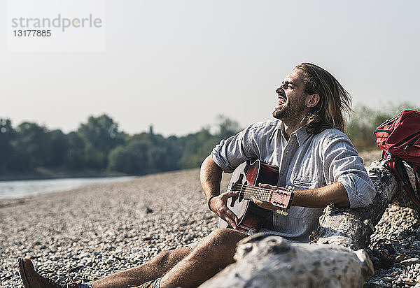 Überschwänglicher junger Mann sitzt am Flussufer und spielt Gitarre
