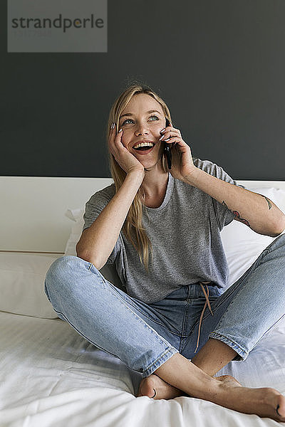 Glückliche junge Frau sitzt im Bett und telefoniert mit dem Handy