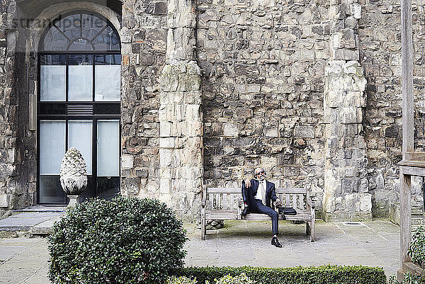 UK  London  älterer Geschäftsmann sitzt auf einer Bank in einem Innenhof und entspannt sich  während er mit Kopfhörern Musik hört