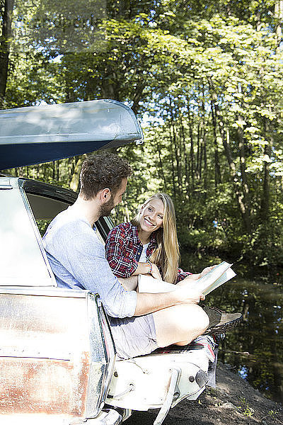 Lächelndes junges Paar mit Karte und Kanu im Auto an einem Bach