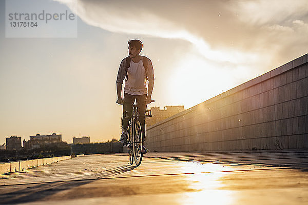 Junger Mann mit Rucksack fährt Fahrrad auf der Promenade bei Sonnenuntergang