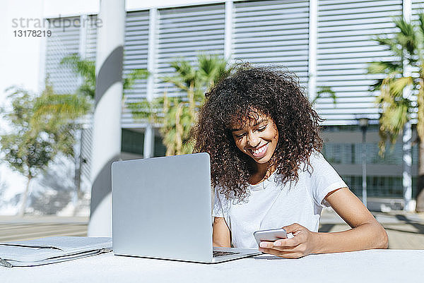 Lächelnde junge Frau mit Laptop  die mit einem Smartphone an einem Tisch im Freien sitzt