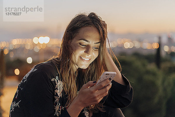 Spanien  Barcelona  Montjuic  lächelnde junge Frau in der Abenddämmerung mit dem Handy