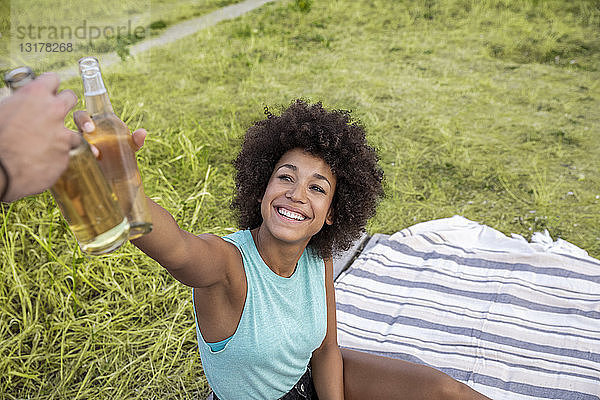 Glückliche Frau sitzt im Freien und klirrt mit einer Bierflasche