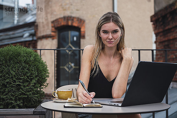 Porträt einer jungen Frau  die mit Laptop auf dem Balkon sitzt und Notizen macht
