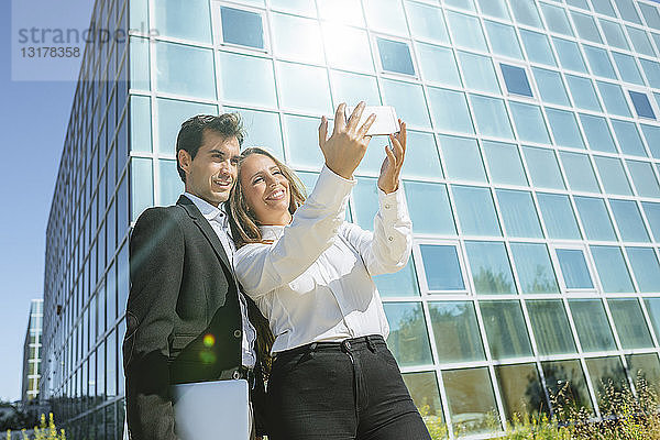 Lächelnde Geschäftsfrau und lächelnder Geschäftsmann beim Selfie vor dem Bürogebäude