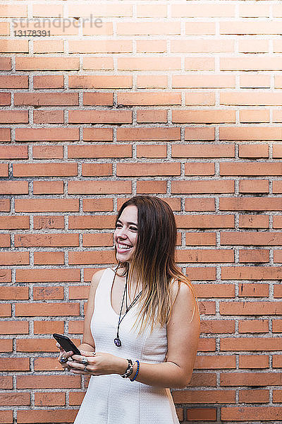 Lachende junge Frau vor einer Ziegelmauer mit Smartphone